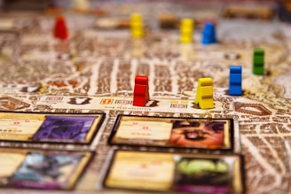 A close-up of a detailed board game in progress with various colored game pieces, such as red, yellow, blue, and green, placed on a textured map. There are also several cards with text and images on them, adding to the intricate and immersive nature of the game.