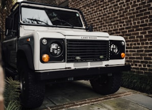 A white Land Rover is parked on a stone driveway, surrounded by brick walls and some greenery visible to the side. The classic design features round headlights and an iconic front grille.