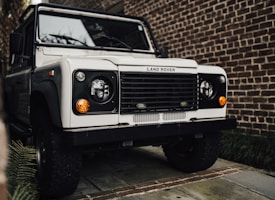 A white Land Rover is parked on a stone driveway, surrounded by brick walls and some greenery visible to the side. The classic design features round headlights and an iconic front grille.