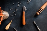 A flat lay of various tools and materials on a dark surface, including a knife with a wooden handle, a small awl, a jar of pencils, rivets, a wooden carving tool, and a metallic pen.