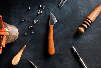 A flat lay of various tools and materials on a dark surface, including a knife with a wooden handle, a small awl, a jar of pencils, rivets, a wooden carving tool, and a metallic pen.