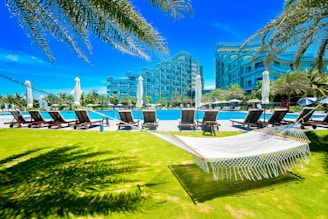 A bright, sunny day at a resort with a luxurious pool area surrounded by lounge chairs and umbrellas. Modern buildings in the background are framed by palm trees. A white hammock hangs in the foreground on a well-manicured green lawn, creating a relaxing atmosphere.