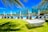 A bright, sunny day at a resort with a luxurious pool area surrounded by lounge chairs and umbrellas. Modern buildings in the background are framed by palm trees. A white hammock hangs in the foreground on a well-manicured green lawn, creating a relaxing atmosphere.