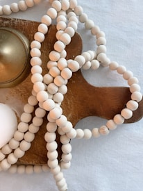 A wooden beaded necklace is laid out on a wooden cutting board. The board has a round brass-colored object next to the beads. The entire arrangement is set against a light-textured background.