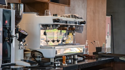 A professional espresso machine sits on a countertop in a café or kitchen setting. Various tools and utensils are nearby, including a coffee tamper and cleaning supplies. The area is well-organized and the machine is a shiny, polished metal.