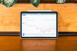 A tablet displaying a Bitcoin to USD chart with fluctuating lines against a wooden surface. Above the chart are navigation tabs including Overview and Market. The setting includes a potted plant with green leaves at the top left of the image.