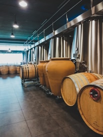 A room filled with large metal tanks and wooden barrels, typically used for winemaking or brewing. The tanks are aligned against one wall under a ceiling with hanging lights, and the barrels are placed in rows along the floor.