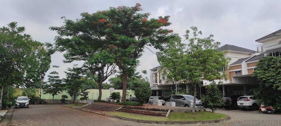 A residential area featuring several modern houses with sloped roofs. The area is landscaped with large, leafy trees, including one with red flowers near the center. Cars are parked in driveways, and the street is paved with bricks, curving along the front of the properties.