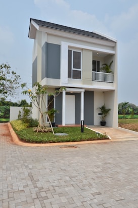 A modern two-story house with a clean architectural design, featuring large windows and a small balcony. The building is surrounded by a neatly landscaped area, including a small tree and green shrubs. The driveway is paved with gray bricks and leads to the front entrance.