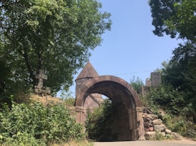 An old stone archway, likely part of a historical or religious site, is surrounded by dense green foliage. A small, pointed structure resembling a roof is visible behind the arch. The archway is constructed with a reddish-brown stone, and there are intricate stone carvings visible nearby.