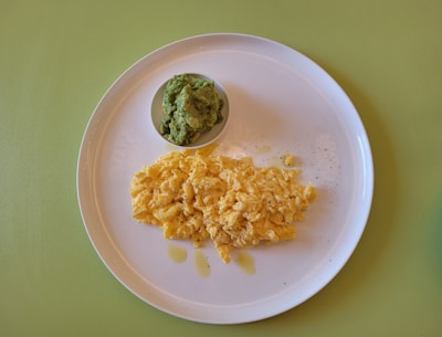 A white plate contains a serving of scrambled eggs with a small bowl of guacamole on a light green surface. The eggs are seasoned with black pepper and appear fluffy and slightly shiny, suggesting they might be freshly prepared.