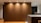 A modern interior space featuring a large wooden wall panel illuminated by recessed ceiling lights. The area includes a kitchen counter on the right with visible modern appliances and shelves with books and kitchen items. The flooring is wooden with a warm tone.