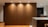A modern interior space featuring a large wooden wall panel illuminated by recessed ceiling lights. The area includes a kitchen counter on the right with visible modern appliances and shelves with books and kitchen items. The flooring is wooden with a warm tone.