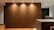 A modern interior space featuring a large wooden wall panel illuminated by recessed ceiling lights. The area includes a kitchen counter on the right with visible modern appliances and shelves with books and kitchen items. The flooring is wooden with a warm tone.