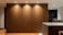 A modern interior space featuring a large wooden wall panel illuminated by recessed ceiling lights. The area includes a kitchen counter on the right with visible modern appliances and shelves with books and kitchen items. The flooring is wooden with a warm tone.