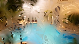 A serene swimming pool is viewed through a frosted glass panel with leaf patterns. The pool water is calm and blue, surrounded by greenery and stone architecture. Sunlight casts gentle shadows, adding a relaxing ambiance.
