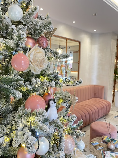 A decorated Christmas tree adorned with pink and white baubles, a faux snow effect, and a small penguin ornament. Beside the tree is a stylish, plush pink sofa and a small round table with magazines. The room has a modern and elegant look with subtle lighting.