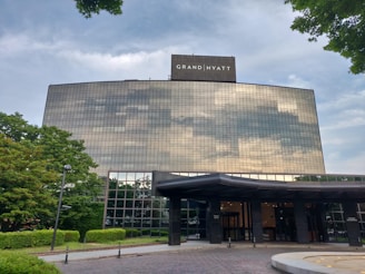 A large, modern hotel building with reflective glass windows that mirror the cloudy sky. The structure is surrounded by well-maintained greenery, including trees and shrubs. The entrance includes a canopy and is set on a paved area.