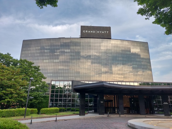 A large, modern hotel building with reflective glass windows that mirror the cloudy sky. The structure is surrounded by well-maintained greenery, including trees and shrubs. The entrance includes a canopy and is set on a paved area.