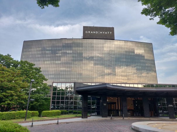 A large, modern hotel building with reflective glass windows that mirror the cloudy sky. The structure is surrounded by well-maintained greenery, including trees and shrubs. The entrance includes a canopy and is set on a paved area.