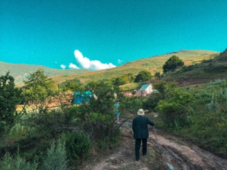 An elderly man wearing a hat walks along a dirt path surrounded by lush greenery and trees. Two small structures with blue and other colored roofs are situated in the background against a backdrop of rolling hills and a vivid blue sky with white clouds.