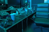 A music studio setting with a mixing console, large speakers, and a laptop displaying audio software. There is a comfortable chair, several beverage bottles, a computer mouse, and headphones lying on the desk. The lighting casts a bluish hue over the scene, giving it a calm, focused ambiance.