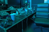 A music studio setting with a mixing console, large speakers, and a laptop displaying audio software. There is a comfortable chair, several beverage bottles, a computer mouse, and headphones lying on the desk. The lighting casts a bluish hue over the scene, giving it a calm, focused ambiance.