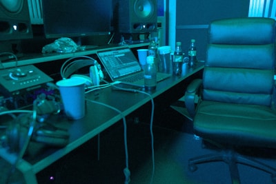A music studio setting with a mixing console, large speakers, and a laptop displaying audio software. There is a comfortable chair, several beverage bottles, a computer mouse, and headphones lying on the desk. The lighting casts a bluish hue over the scene, giving it a calm, focused ambiance.