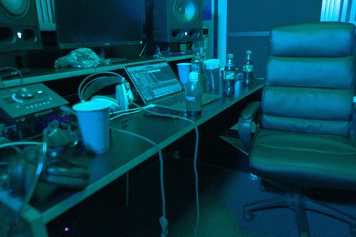 A music studio setting with a mixing console, large speakers, and a laptop displaying audio software. There is a comfortable chair, several beverage bottles, a computer mouse, and headphones lying on the desk. The lighting casts a bluish hue over the scene, giving it a calm, focused ambiance.