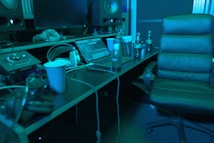 A music studio setting with a mixing console, large speakers, and a laptop displaying audio software. There is a comfortable chair, several beverage bottles, a computer mouse, and headphones lying on the desk. The lighting casts a bluish hue over the scene, giving it a calm, focused ambiance.