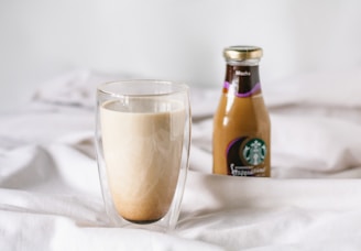 A clear double-walled glass filled with a creamy coffee beverage is placed on a soft, white fabric background. Beside it, there is a bottled mocha frappuccino with a gold cap, featuring a recognizable coffee brand's logo.