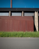A large industrial building with a rust-colored facade stands behind a row of tall green plants, set against a clear blue sky. The building features a tall chimney and a series of metal grates or vents near the roof.