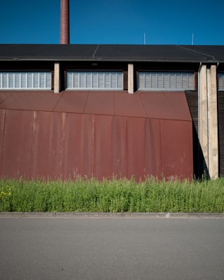 A large industrial building with a rust-colored facade stands behind a row of tall green plants, set against a clear blue sky. The building features a tall chimney and a series of metal grates or vents near the roof.