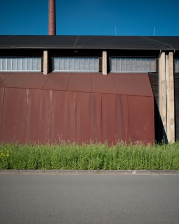 A large industrial building with a rust-colored facade stands behind a row of tall green plants, set against a clear blue sky. The building features a tall chimney and a series of metal grates or vents near the roof.