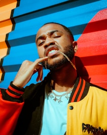 A person poses against a colorful, striped mural with bold blue, red, and orange sections. They are wearing a varsity-style jacket with red, black, and yellow colors, a white shirt, and several necklaces, including one chain in their mouth.
