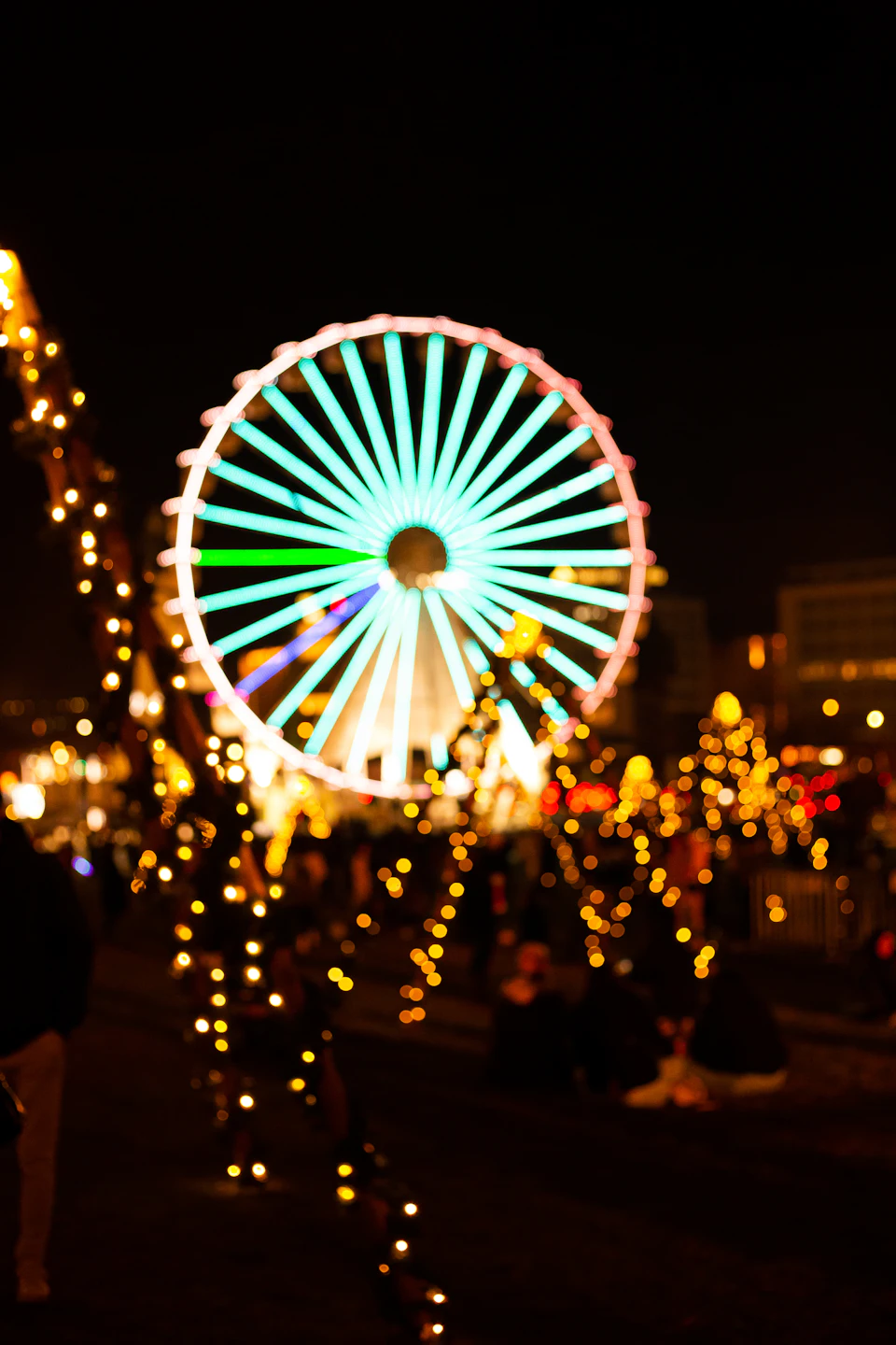 The image depicts a large, illuminated Ferris wheel at night, with its blue and white colors standing out against the dark sky. The ferris wheel is surrounded by numerous lights that twinkle like stars, creating a festive atmosphere. In the foreground, there are people gathered around the ferris wheel, adding to the lively scene.