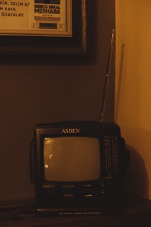 A vintage ASBEN mini personal television with an AM/FM radio feature sits on a wooden surface. It has a small screen and various dials and buttons for volume and tuning. An antenna is extended from the top of the television. Above the television, a framed theater or event poster is partially visible.