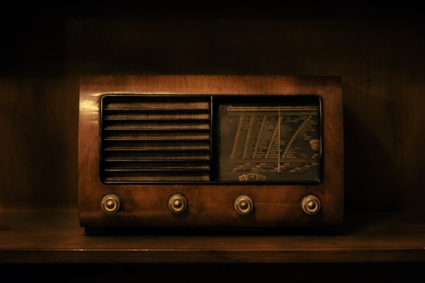 A vintage radio with a wooden exterior, featuring four knobs on the front and a combination of slatted speaker grille with a dial tuning display. The image is dimly lit and reflects an antique aesthetic.