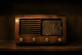 A vintage radio with a wooden exterior, featuring four knobs on the front and a combination of slatted speaker grille with a dial tuning display. The image is dimly lit and reflects an antique aesthetic.