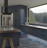 A modern kitchen interior features sleek and dark cabinetry with a glossy refrigerator. The countertop holds a contemporary ceramic vase with a green plant near a large window offering a view of trees. An island with a black stove and ventilation hood enhances the sophisticated look, and a textured rug covers part of the wooden floor.