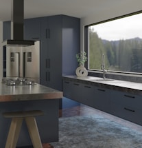 A modern kitchen interior features sleek and dark cabinetry with a glossy refrigerator. The countertop holds a contemporary ceramic vase with a green plant near a large window offering a view of trees. An island with a black stove and ventilation hood enhances the sophisticated look, and a textured rug covers part of the wooden floor.