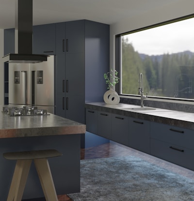 A modern kitchen interior features sleek and dark cabinetry with a glossy refrigerator. The countertop holds a contemporary ceramic vase with a green plant near a large window offering a view of trees. An island with a black stove and ventilation hood enhances the sophisticated look, and a textured rug covers part of the wooden floor.