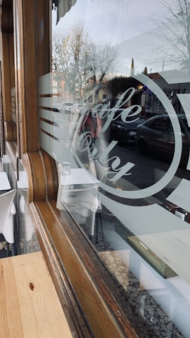 A cozy cafe setting with wooden accents and a large glass window displaying the words 'Cafe Only'. The reflection shows a street view with parked cars and leafless trees, indicating a cold season. Inside, there are white chairs and tables set up for customers.