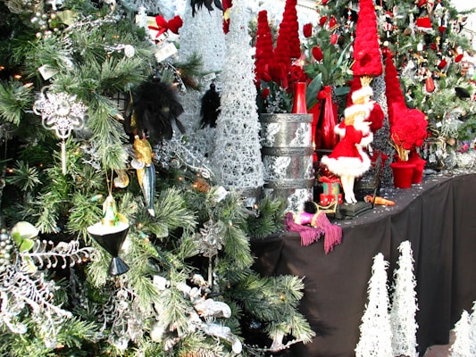 A festive display featuring decorated Christmas trees adorned with ornaments and baubles. There are red and white fur-trimmed decorative elements resembling Santa figures and various red and silver decorative objects are placed on a table covered with a black cloth. The scene is filled with green pine tree branches, red flowers, and shiny white decor.