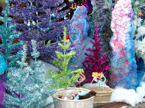 A colorful and eclectic display featuring an assortment of artificial trees in vibrant hues of blue, purple, green, and pink. The trees are adorned with shiny tinsel and beads. In the foreground, a wicker basket contains various objects, including a small doll and pinecones. Nearby, whimsical statues of mermaids add to the fantastical theme, while a collection of textured objects resembling large, colorful rocks or corals provides an imaginative backdrop.