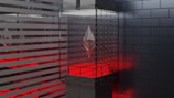 A modern and futuristic display setup featuring a clear glass case containing a geometric metallic symbol that resembles the Ethereum logo. The surrounding environment is designed with sleek, horizontal gray stripes and a dark-tiled wall, illuminated by red ambient lighting.