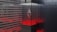 A modern and futuristic display setup featuring a clear glass case containing a geometric metallic symbol that resembles the Ethereum logo. The surrounding environment is designed with sleek, horizontal gray stripes and a dark-tiled wall, illuminated by red ambient lighting.