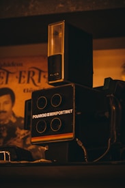 A vintage Polaroid camera is placed on a shelf with an orange-tinted photograph in the background. The camera is labeled 'Polaroid Miniportrait' and features four lens openings. Overhead lighting casts a warm glow, contributing to an overall nostalgic atmosphere.