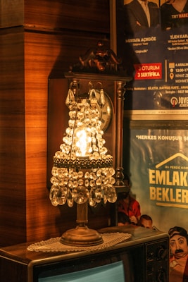 A vintage-style table lamp with crystal ornaments emits a warm glow in a retro setting. The lamp rests on a wooden surface, possibly an old television, and is surrounded by wooden paneling. In the background, there is a wall decorated with posters featuring people and text, adding a nostalgic touch to the scene.