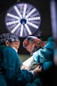 Two surgeons wearing masks and colorful caps are intensely focused on performing surgery under a bright surgical light. They are dressed in teal surgical gowns and white gloves, with a variety of surgical instruments in use.